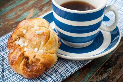 Swedish cinnamon berry bun served on a rustic wooden table with a cup of freshly brewed coffee in a blue and white striped cup and saucer. Coffee and a bun is a tradition in Sweden known as fika.