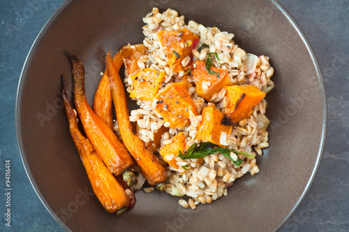 Roasted butternut squash, shallots and carrots with spelt risotto. Served in a rustic brown bowl on a blue slate background.