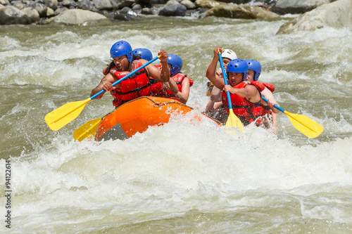 A team in an orange raft navigate choppy whitewater rapids in Ecuador, paddling with yellow oars while wearing blue helmets and red life jackets amid rushing water and rocky riverbanks.