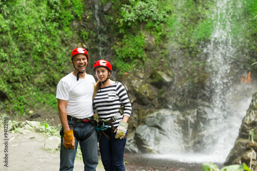 A man and woman in red helmets stand together smiling in front of a lush green waterfall with moss-covered rocks near Banos, Ecuador, a vibrant adventurous moment.