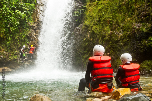 People in red life jackets and white helmets sit on rocks watching a cascading waterfall in a lush green canyon near Banos, Ecuador, mist rising from the water during a canyoning adventure.
