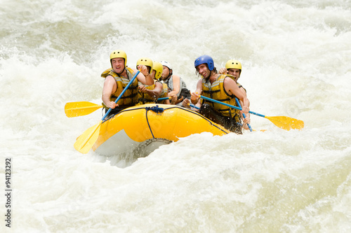 A group of tourists in helmets and life jackets navigate through the white water rapids of a river in Ecuador on an extreme rafting adventure with a guide.