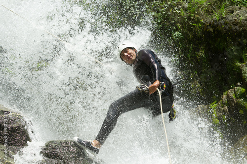 Fotografie A man in black neoprene gear rappels down a cascading waterfall gripping a rope in the lush green Chama Canyon of Ecuador, splashing white water around him in a thrilling canyoning descent