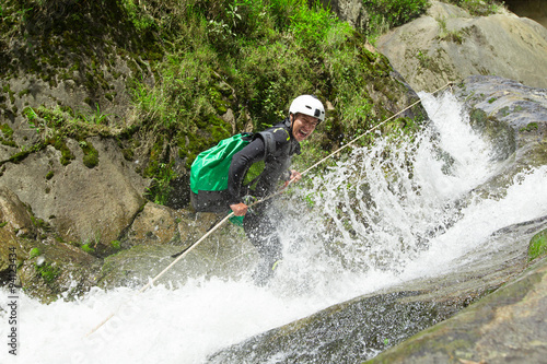 A backpacker in a white helmet and green backpack rappels down a fast-flowing canyon waterfall surrounded by lush green mossy rocks in Ecuador, descending toward the river in a rugged canyoning sport.