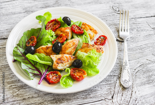Fresh salad with chicken breast, sun-dried tomatoes, green salad and olives on a white plate on a wooden surface. Healthy food