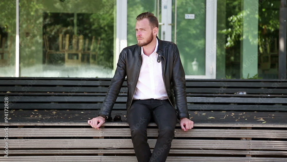 A man 29 years old sitting down outside on a bench, wearing a white shirt with a black leather jacket and jeans, thinking on a sunny summer day. A man 29 years old sitting down outside on a bench, wearing a white shirt with a black leather jacket and jeans, thinking on a sunny summer day.