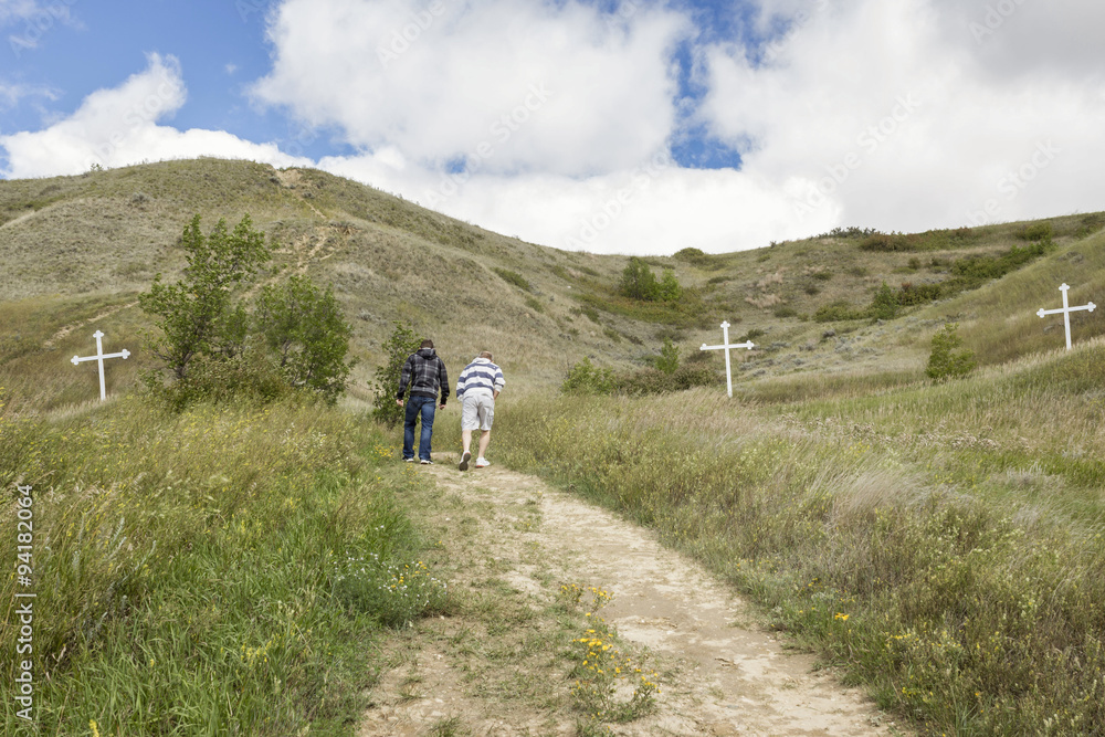 horizontal image of a father and son walking on a path up a steep hill with white crosses on either side of hill under a blue sky with white cloud in the summer time.