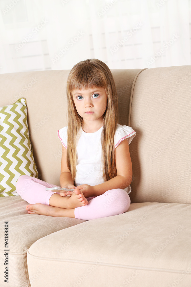 Beautiful little girl with smart phone sitting on sofa, on home interior background Beautiful little girl with smart phone sitting on sofa, on home interior background