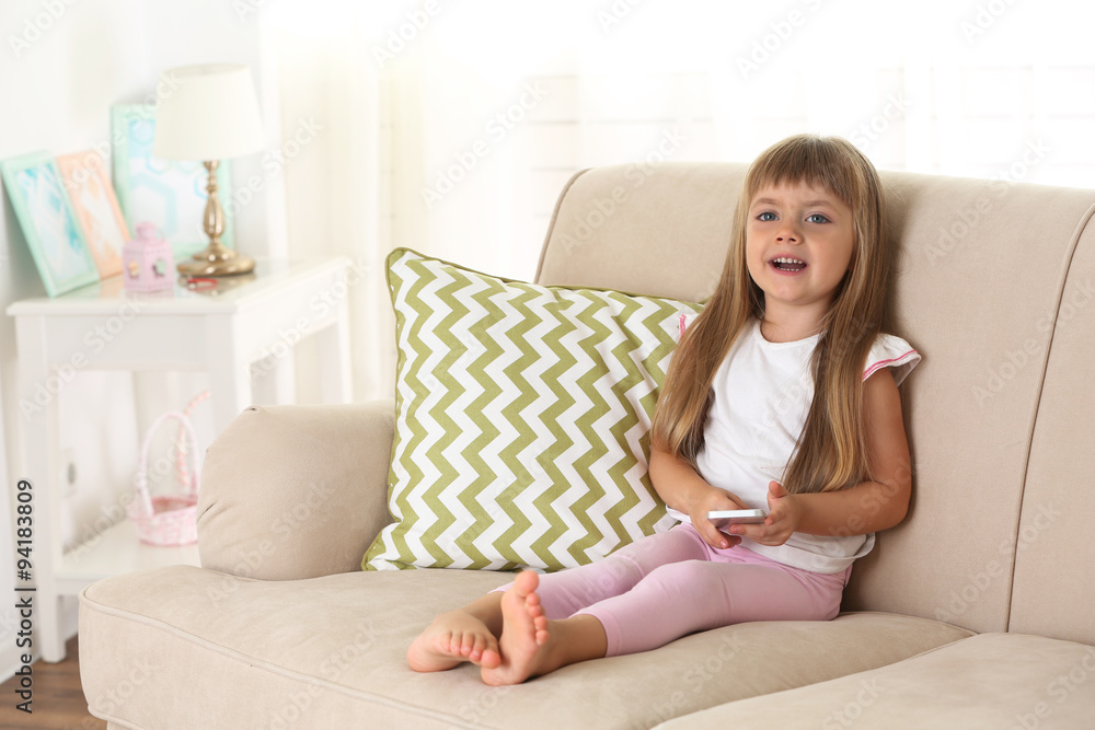 Beautiful little girl with smart phone sitting on sofa, on home interior background Beautiful little girl with smart phone sitting on sofa, on home interior background