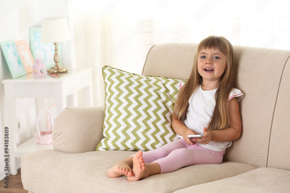 Beautiful little girl  with smart phone sitting on sofa, on home interior background