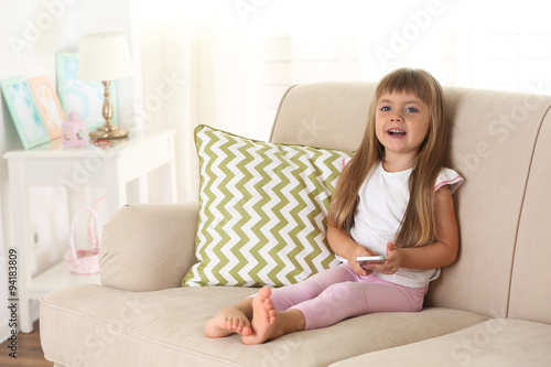 Beautiful little girl with smart phone sitting on sofa, on home interior background