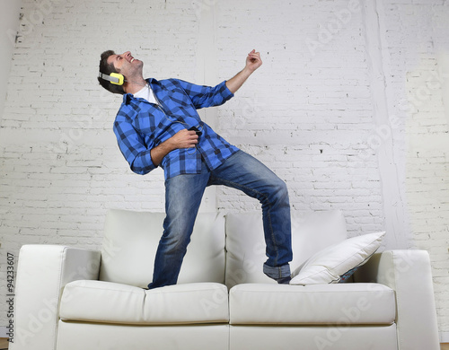 20s or 30s man jumped on couch listening to music on mobile phone with headphones playing air guitar photo