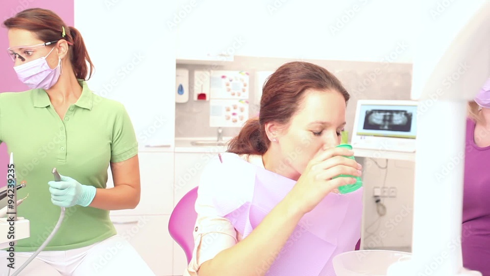 Female datient drinking water and sitting in a pink dental chair while female dentist preparing tools / camera moving left Female datient drinking water and sitting in a pink dental chair while female dentist preparing tools / camera moving left
