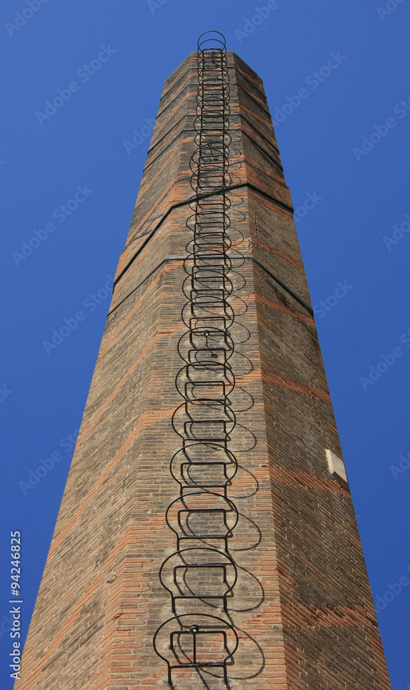 Old red brick chimney (low angle shot) with weathered brickwork & ladder against a blue sky