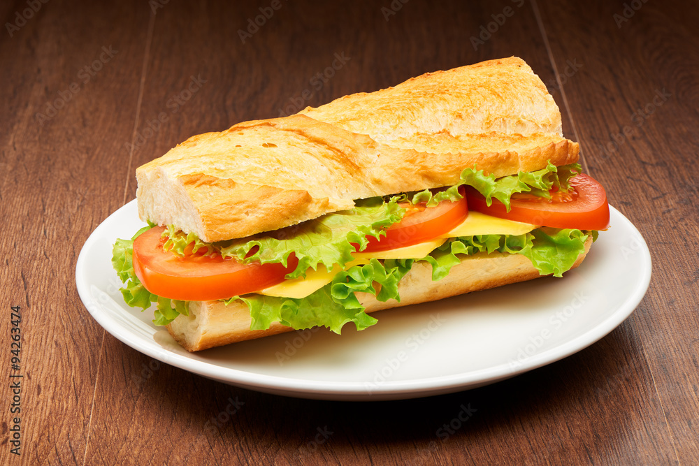 Tomato, cheese and salad sandwich from fresh baguette on white ceramic plate on dark wooden table background