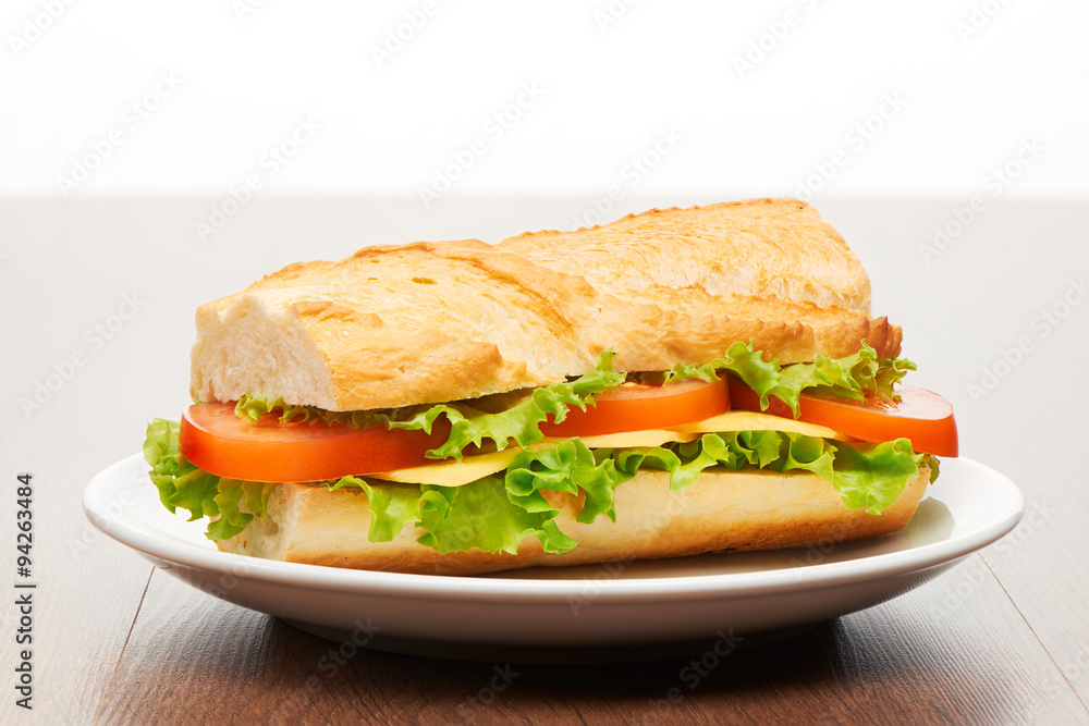 Tomato, cheese and salad sandwich from fresh baguette on white ceramic plate on bright light brown wooden table background Tomato, cheese and salad sandwich from fresh baguette on white ceramic plate on bright light brown wooden table background