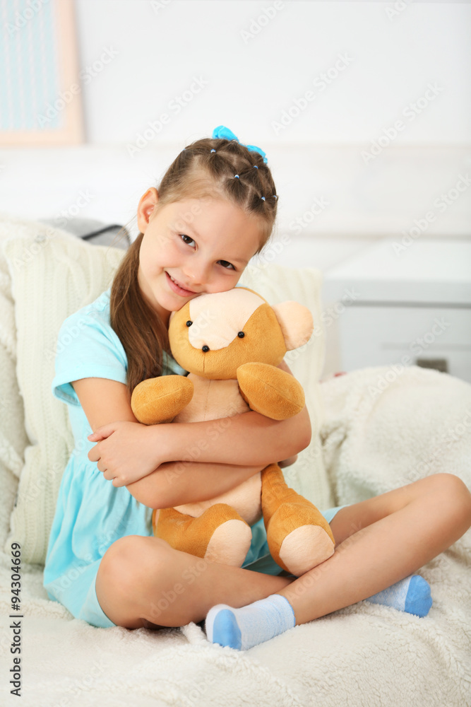 Beautiful little girl with teddy bear sitting on sofa, on home interior background Beautiful little girl with teddy bear sitting on sofa, on home interior background