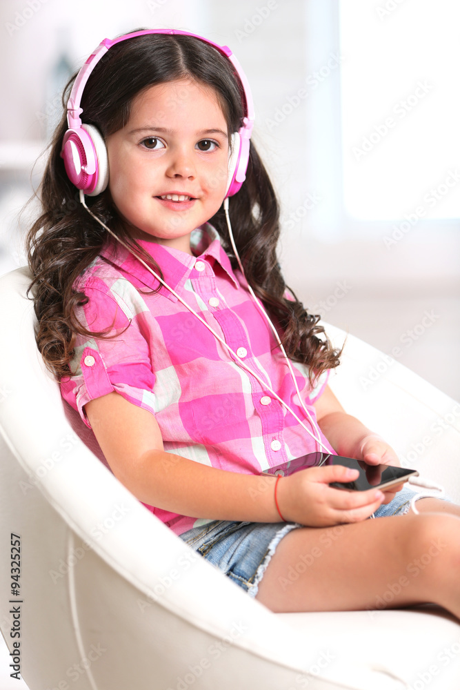 Attractive little girl sitting on comfortable chair and listening music with pink headphones in the room, close up Attractive little girl sitting on comfortable chair and listening music with pink headphones in the room, close up