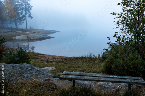 old bench on the shore by a lake covered in fog an early autumn morning