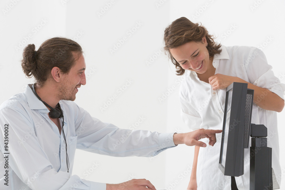 Obraz premium Two young colleagues, a boy sitting and a girl bowing next to his desk, talking and pointing to a computer screen
