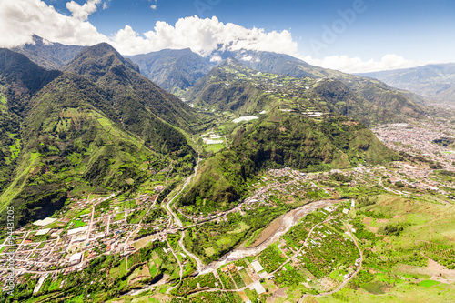 An aerial view of Banos de Agua Santa in a lush green terraced valley with winding roads and the Pastaza River below the Tungurahua volcano in the Andes of Ecuador.