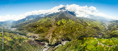 A breathtaking aerial panorama of vibrant green mountain slopes and the snow-capped Tungurahua volcano rising above white clouds near Banos, Ecuador, rugged terrain under a clear blue sky.