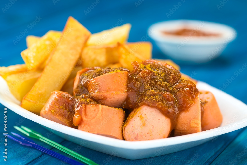 German fast food called Currywurst served with French fries on a disposable plate with party forks in the front (Selective Focus, Focus on the front of the sausage on top) German fast food called Currywurst served with French fries on a disposable plate with party forks in the front (Selective Focus, Focus on the front of the sausage on top)