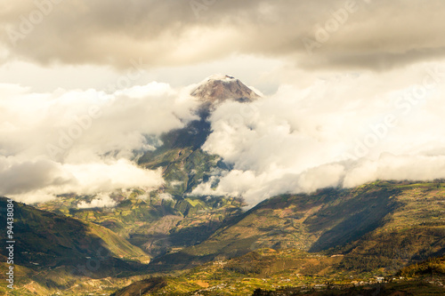 A majestic mountain peak rises above a thick layer of clouds with lush green slopes and shadowed valleys below near Banos, Ecuador, a serene scene of natural grandeur in soft diffused light.