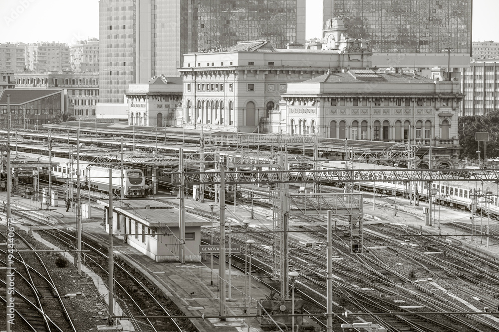 Obraz premium A monochrome view of the busy Genova Brignole railway station in Italy, numerous intersecting tracks and overhead lines before a mix of historic and modern buildings, conveying industrial city life.