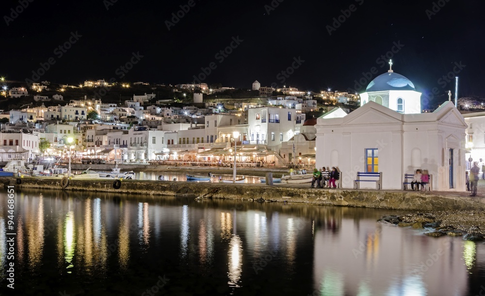 Night view of Chora port in Mykonos,Greece.Hora town cityscape lights reflected on sea,whitewashed blue dome greek island church in the harbour and yachts anchored. A colourful and beautiful seascape.