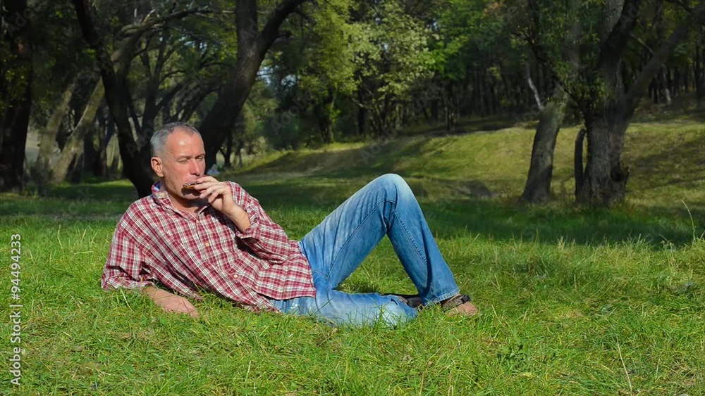 Active man resting lying on a green meadow and plays the harmonica