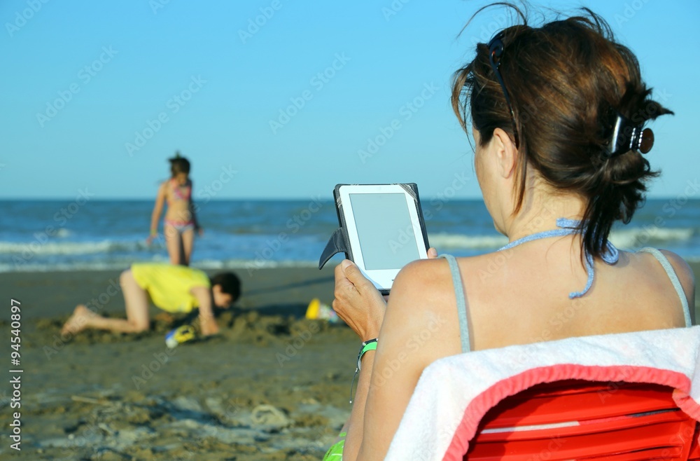 woman reads the ebook on the beach while her children playing