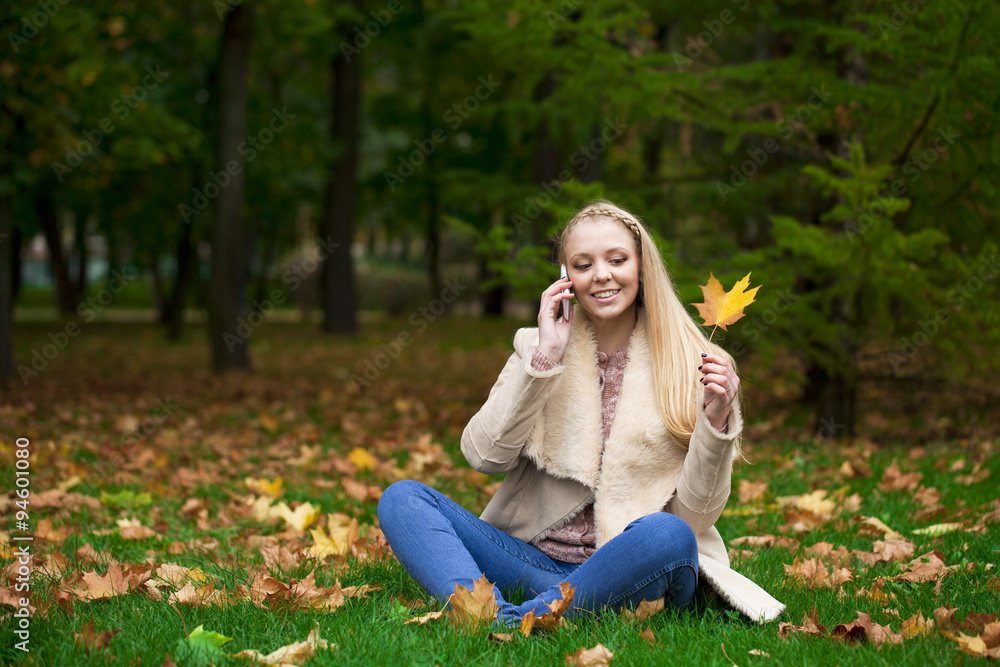 Naklejka premium Young happy blonde woman calling by phone in autumn park