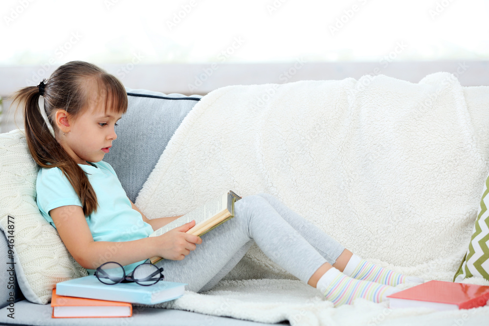 Beautiful little girl sitting on sofa with book, on home interior background Beautiful little girl sitting on sofa with book, on home interior background