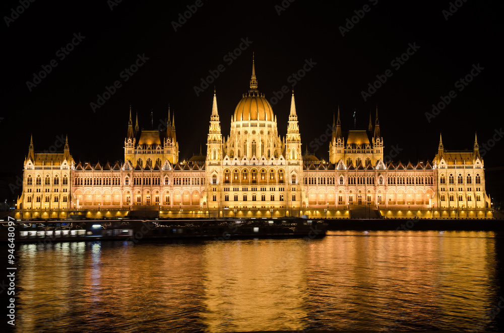 Night view of the famous Parliament building of Hungary in Budapest, illuminated by electric light. The reflections of electric lighting in the waters of the Danube River. 