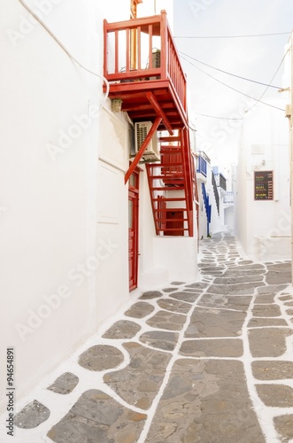 A very traditional alley view of the architecture in Chora, on the greek island Mykonos, Greece. A red door, balcony, fence and windows of a whitewashed house.