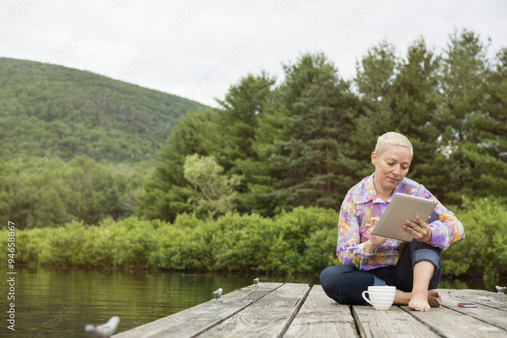 A woman sitting outdoors on a jetty using a digital tablet. A woman sitting outdoors on a jetty using a digital tablet.
