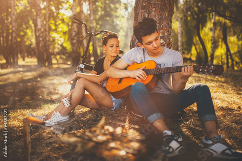 Teen boy sitting with his girlfriend by a tree, having a picnic and playing guitar on a sunny autumn day in forest. Teenagers loving couple relaxing by bonfire outdoors