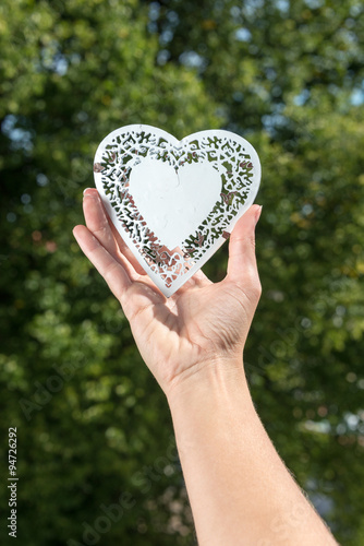 Hand holds up heart made of white metal in a park in front of tr