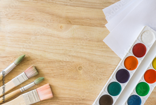 Four painting brushes in the left bottom corner, white paper and watercolor on wooden background