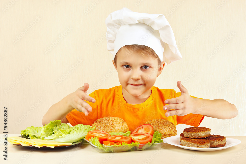 Little boy in chef hat at the table with ingredients for hamburger Little boy in chef hat at the table with ingredients for hamburger
