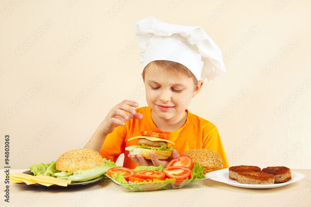 Little smiling boy in chefs hat puts tomato on the hamburger Little smiling boy in chefs hat puts tomato on the hamburger