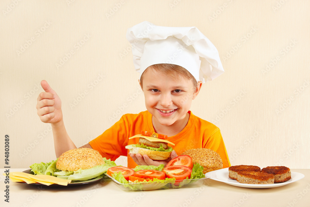 Young funny boy in chefs hat shows how to cook a hamburger Young funny boy in chefs hat shows how to cook a hamburger