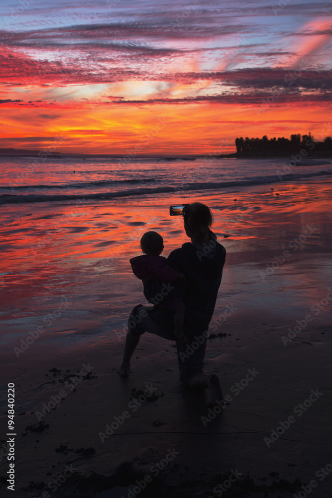 Woman with child takes picture of purple and orange sunset looking towards Anacapa Island, Ventura, California, USA, 12.16.2013