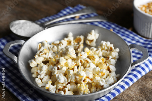 Salted popcorn in a metal bowl with cup of corns on checked cotton napkin on wooden table
