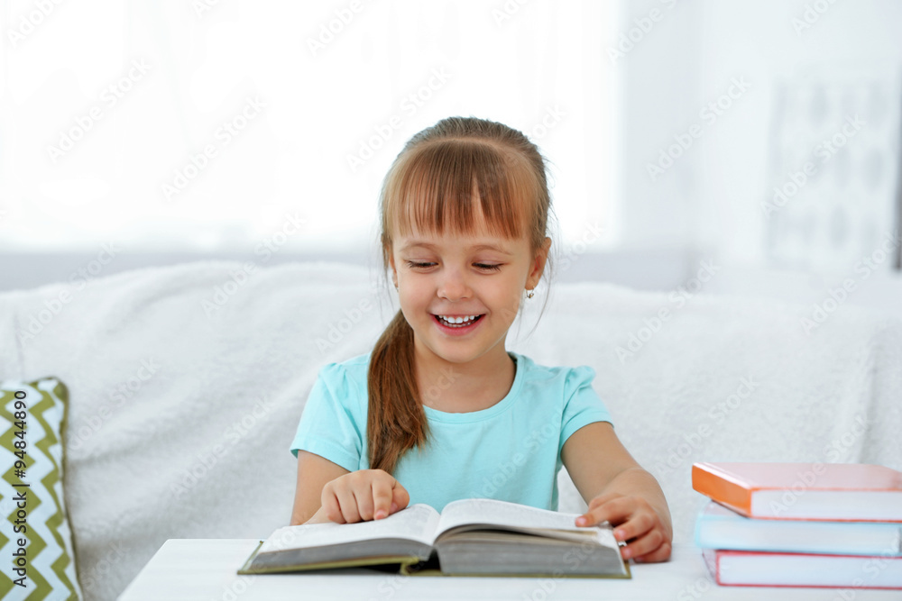 Beautiful little girl sitting on sofa with book, on home interior background Beautiful little girl sitting on sofa with book, on home interior background