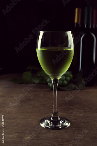 White wine glass against different bottles and grape on wooden table