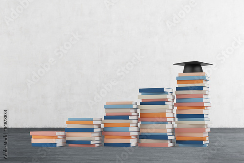 A stair is made of colourful books. A graduation hat is on the final step. Concrete wall and wooden floor.