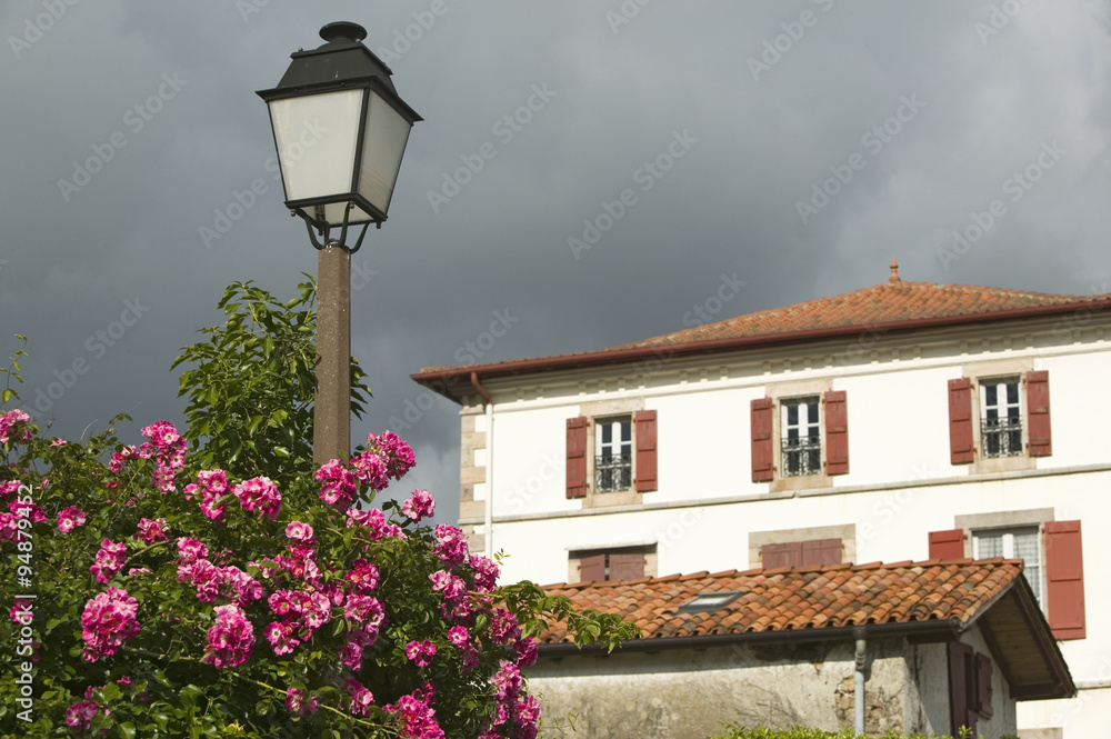Summer flowers, lamp post and home in Sare, France in Basque Country on Spanish-French border, a hilltop 17th century village in the Labourd province. The houses are built in the traditional style of the region, with shutters painted colors red and green of the Basque flag. Close to St. Jean de Luz, on the Cote Basque, France.