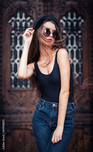 young beautiful brunette girl in a hat  sunglasses  t-shirt and jeans  with a film camera walks in the old city  holding hand hat for fear of losing it